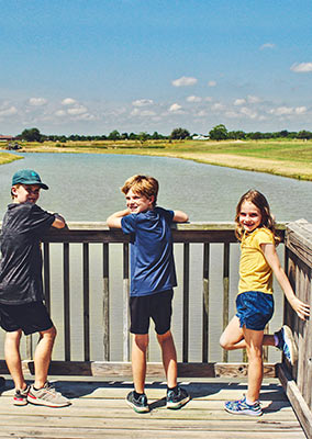 Two young boys and their little sister wearing shorts and tshirts smiling and standing on one of Lake Dewberry RV Resort's docks overlooking the lake under sunny blue skies